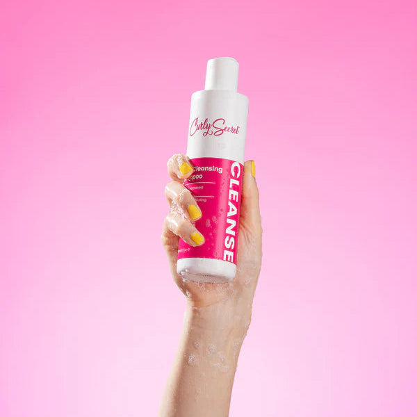 Hand met natte vingers houdt Curly Secret Deep Cleansing Shampoo vast tegen roze achtergrond, ideaal voor diepe reiniging van krullend haar.
