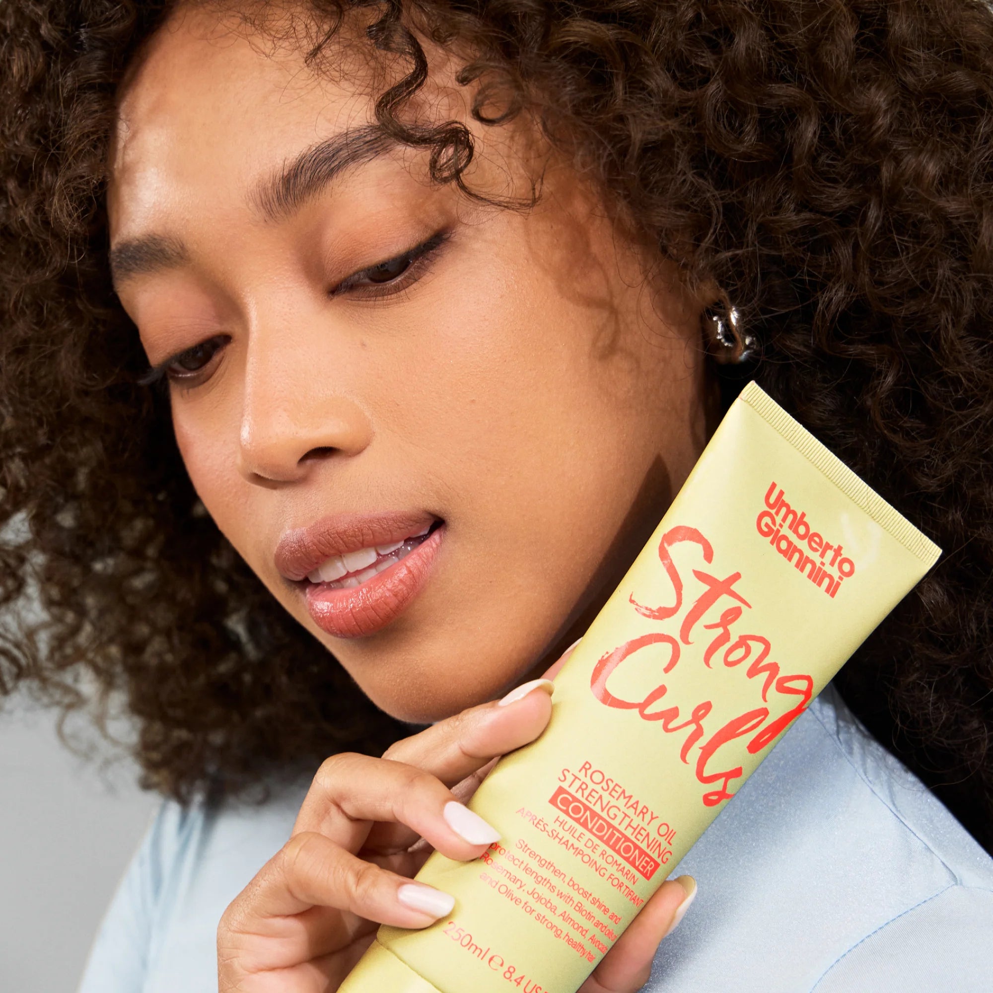 Vrouw met krullend haar toont Umberto Giannini Strong Curls Rosemary Conditioner 250ml, ideaal voor gevoelige hoofdhuid en CG haarverzorging.