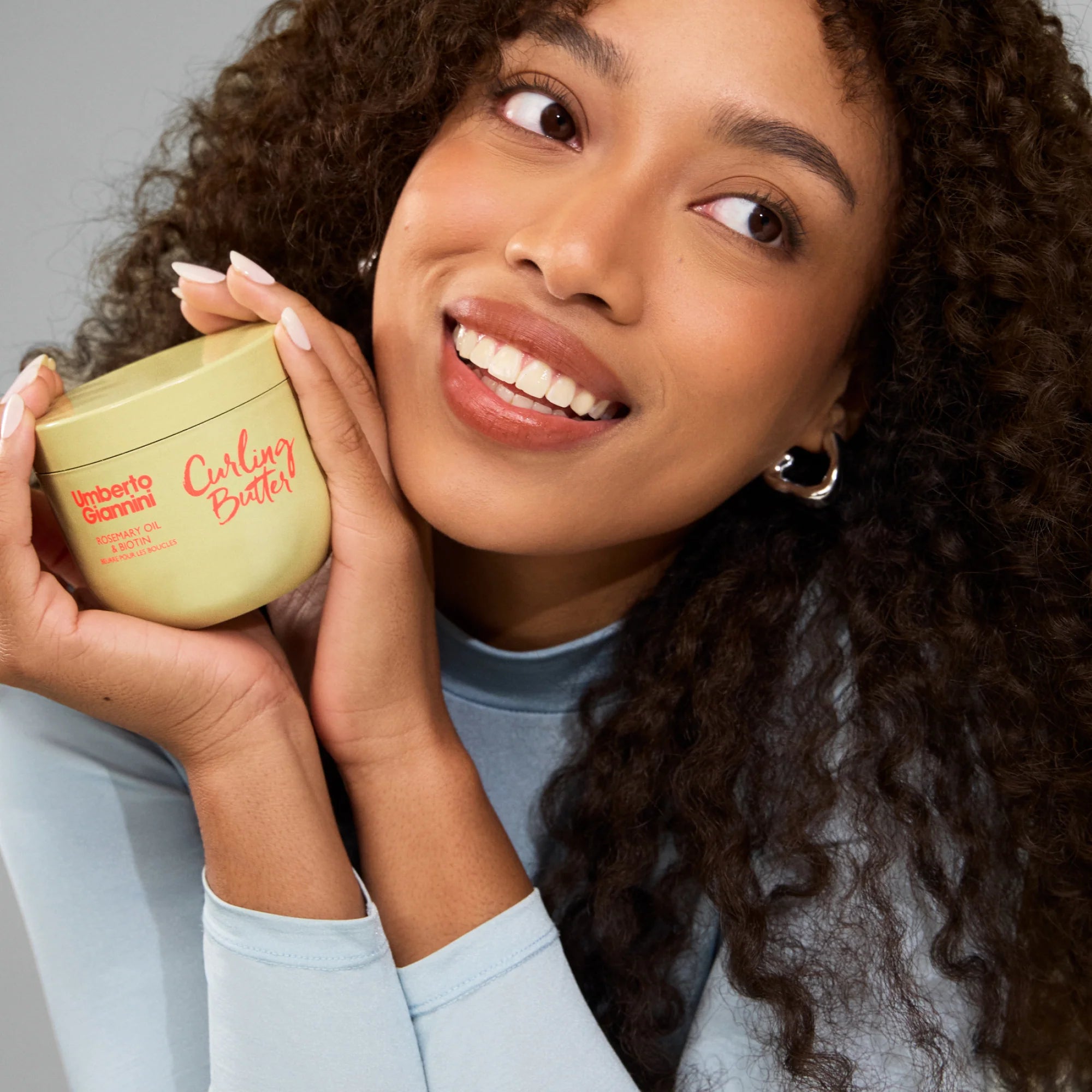 Vrouw met krullend haar houdt Umberto Giannini Strong Curls Curling Butter 300ml vast, ideaal voor styling en verzorging van natuurlijke krullen.
